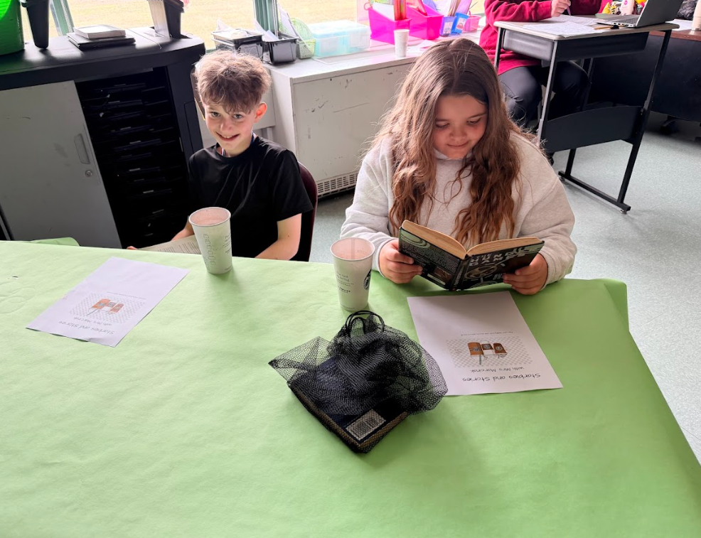 Image of student sitting around a table with a green table cloth, enjoying hot cocoa while reading a book