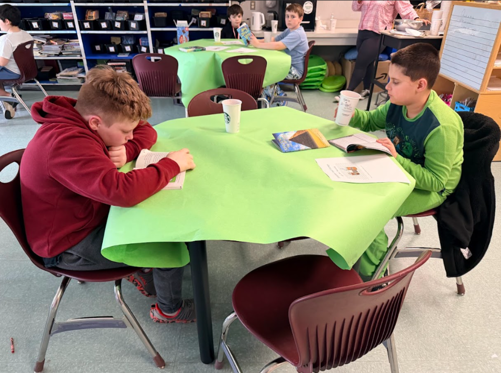 Image of student sitting around a table with a green table cloth, enjoying hot cocoa while reading a book