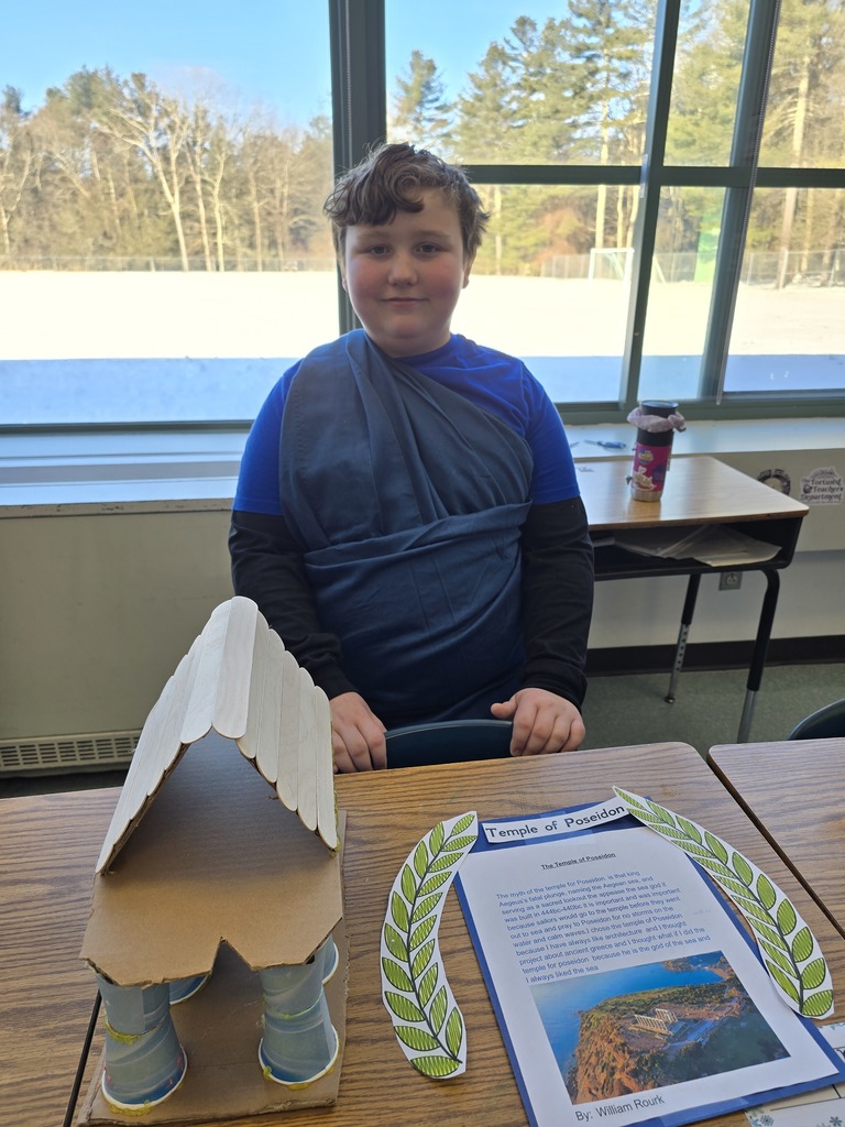 Image of a student in a toga made of a blue bed sheet standing behind his Greek Mythology project about the Temple of Poseidon