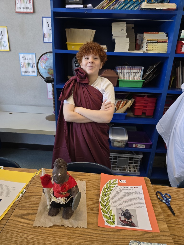 Image of a student in a toga made of a burgundy bed sheet standing behind his Greek Mythology project about Ares