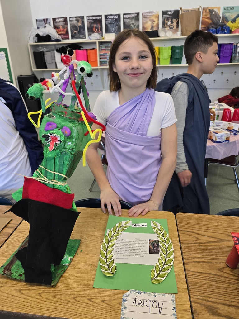 Image of a student in a toga made of a purple bed sheet standing behind her Greek Mythology project about Medusa