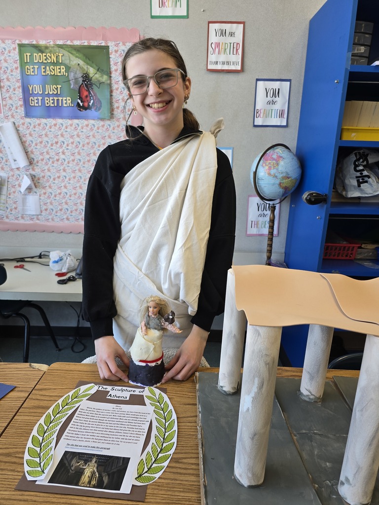 Image of a student in a toga made of a white bed sheet standing behind her Greek Mythology project about Athena