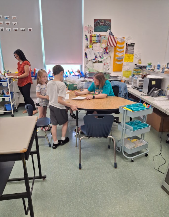 Image of a teacher working with two students at her desk. Another educator stands in the background.