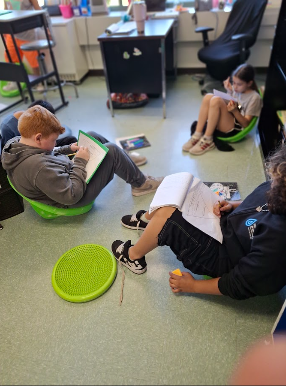 Image of students, sitting in flexible seating on the floor, working on a writing assignment