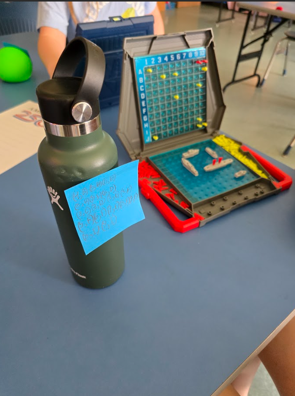 Image of a water green water bottle with a blue sticky note with coordinates on it in front of a game of Battleship
