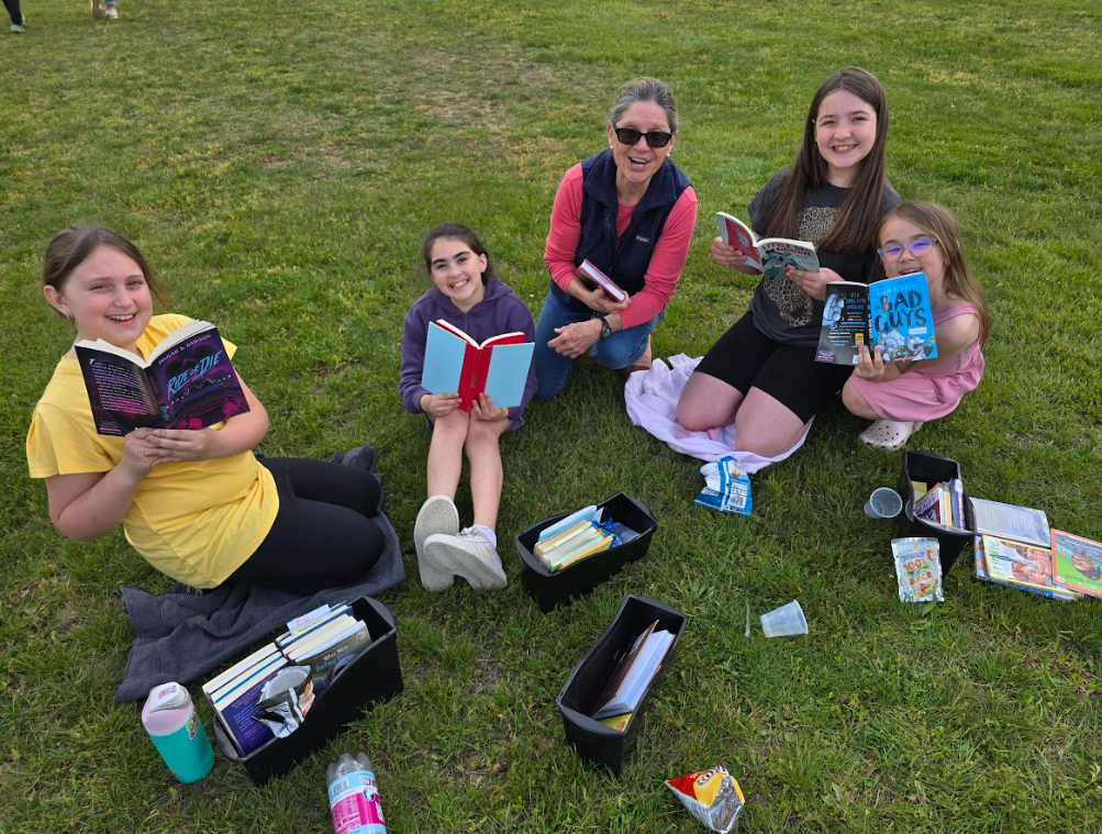 Image of children sitting outside on the grass with with their teacher reading books with a snack