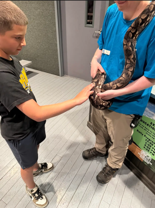 Image of a student petting a snake that is held by an employee of the reptile den