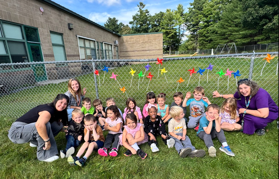 Image of students posing with their teachers in front of the pinwheels they placed in a fence to observe the annual International Day of Peace