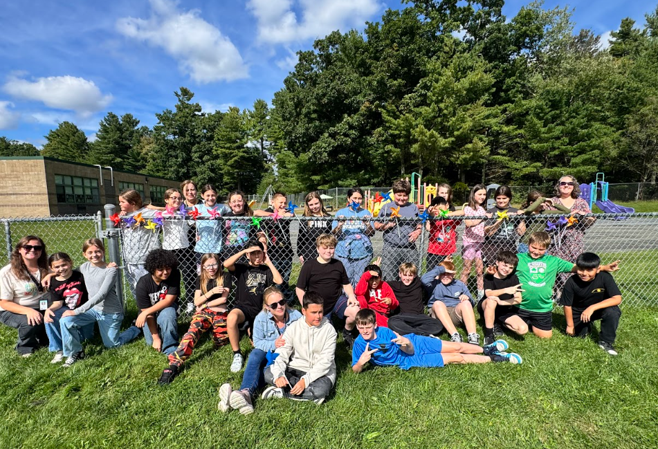 Image of students posing with their teachers in front of the pinwheels they placed in a fence to observe the annual International Day of Peace