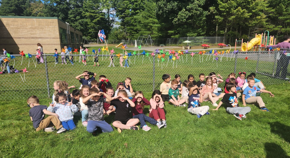 Image of students posing with their teachers in front of the pinwheels they placed in a fence to observe the annual International Day of Peace