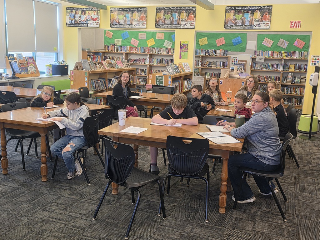 Children & adults sit at tables in a library participating in a meeting