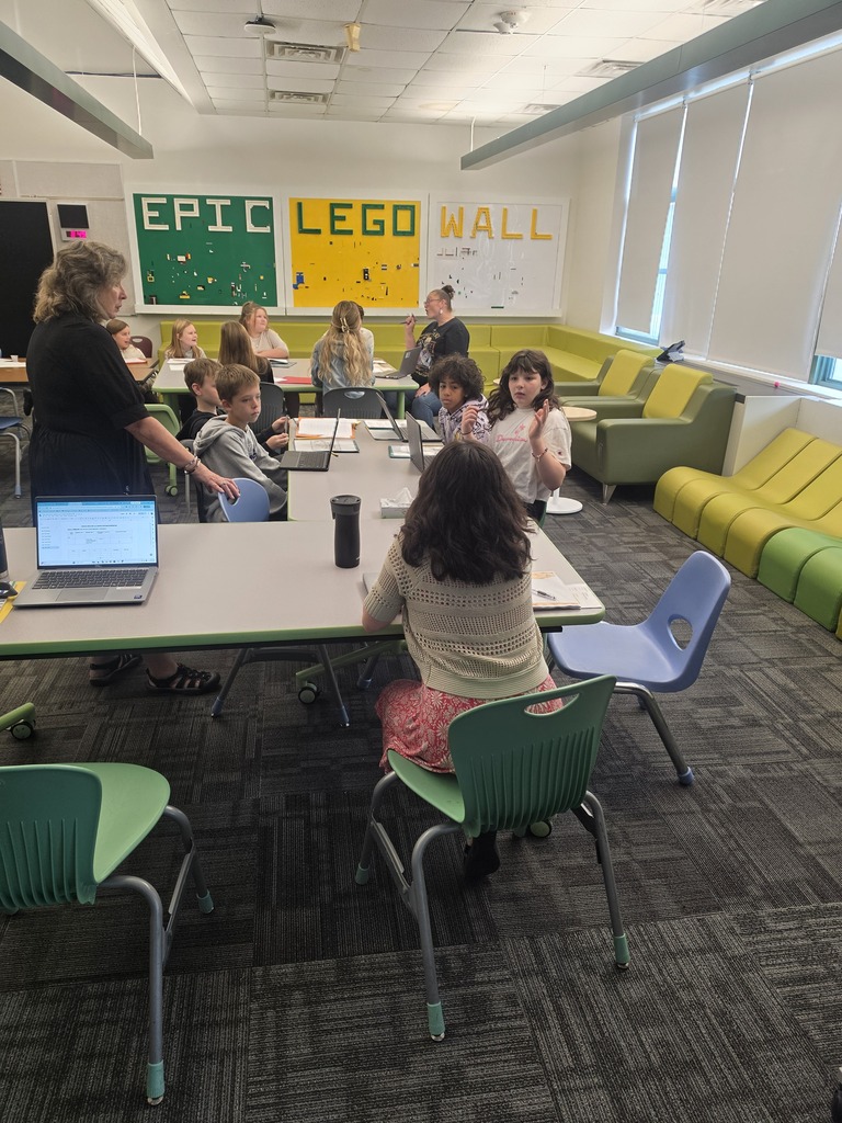 Students and adults sit at or stand around tables participating in a meeting