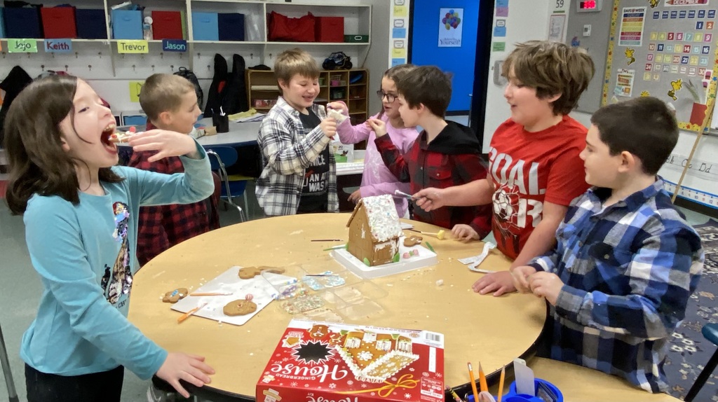 image of children surrounding a table as they work together to construct a gingerbread house while one student pretends to eat a piece of the decorated house.