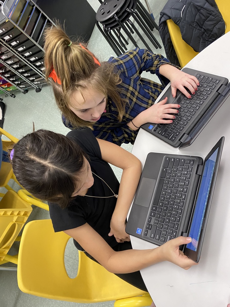 Two female students sit at a desk with their laptops doing research