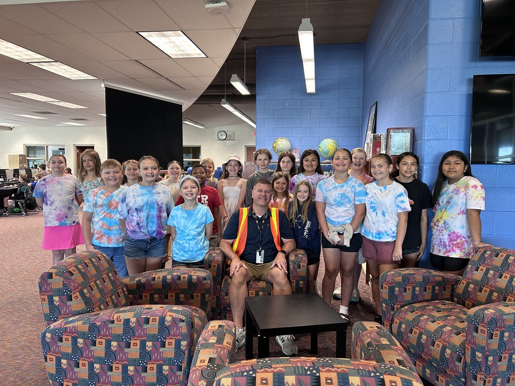 A group of young people and an adult stand and sit in a room with blue walls. Some wear tie-dye shirts.