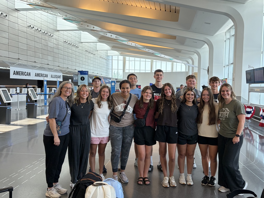 A group of individuals stands together in an airport, wearing casual clothing. Some are smiling and appear to be posing for a photo.
