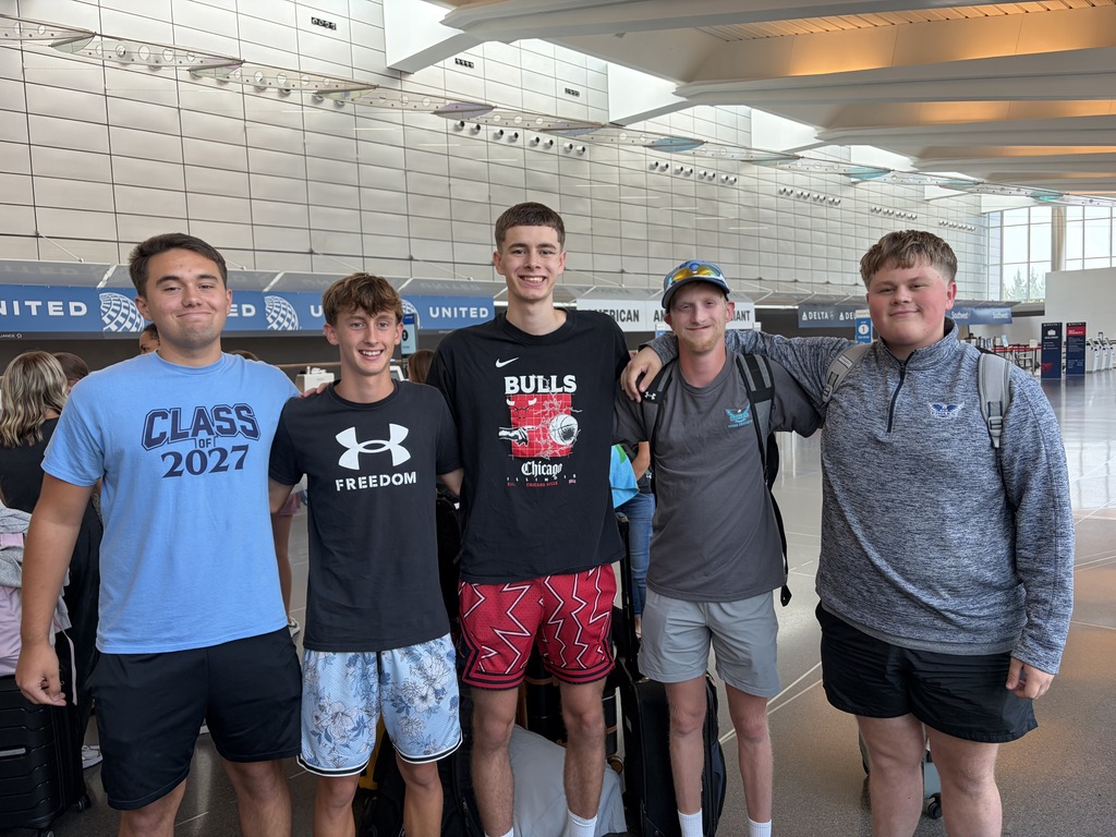 Five young men in casual clothing stand in an airport, smiling. One wears a "CLASS 2027" shirt.