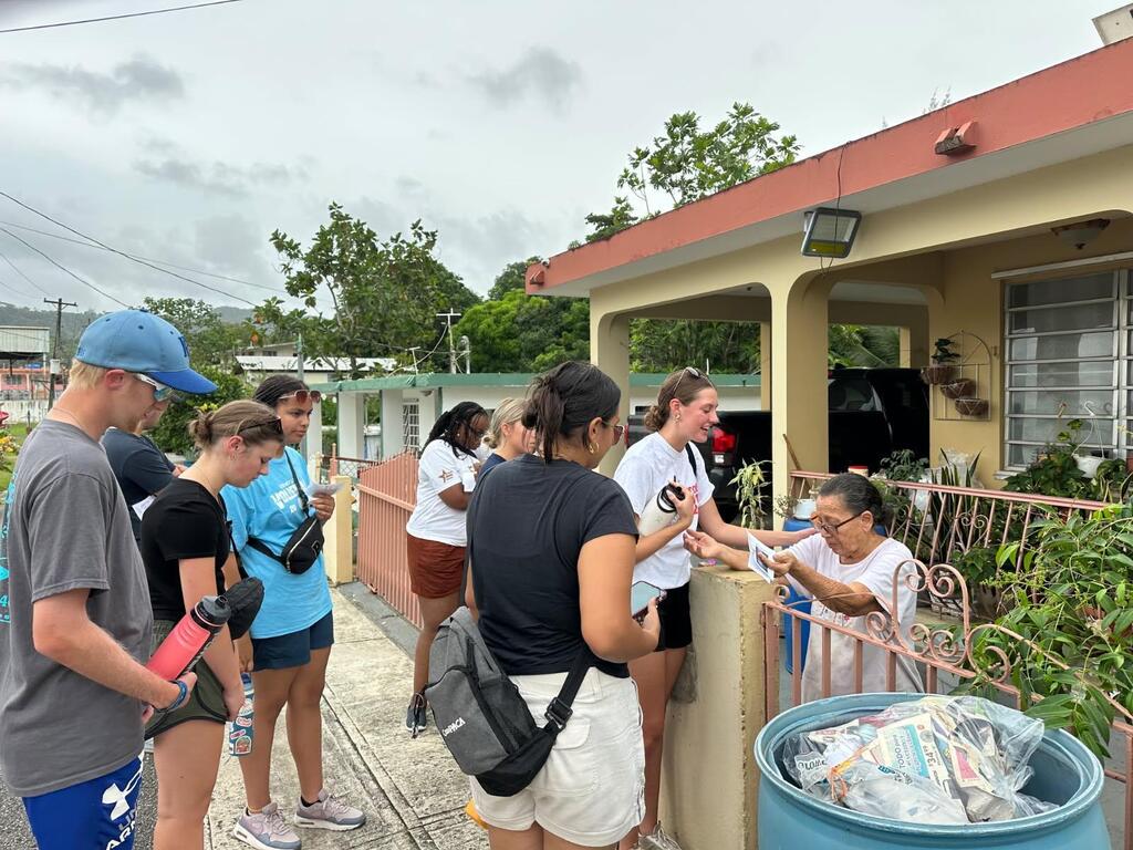 Several people outside a house with a fence, some interacting with a woman. Trees and a trash bin are nearby.