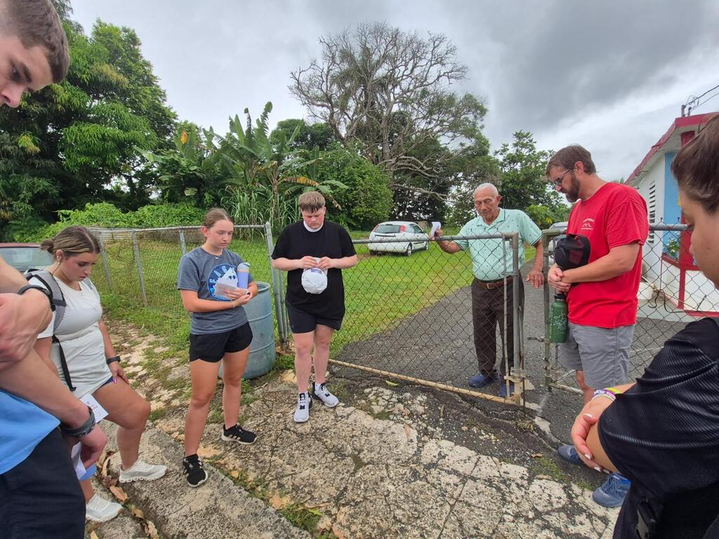 A group of people stands in front of a metal gate. They seem to be engaged in a conversation.