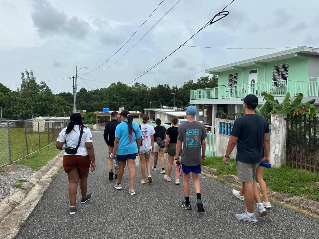 A group of people walking on a paved street with a fence, trees, and a house in the background.