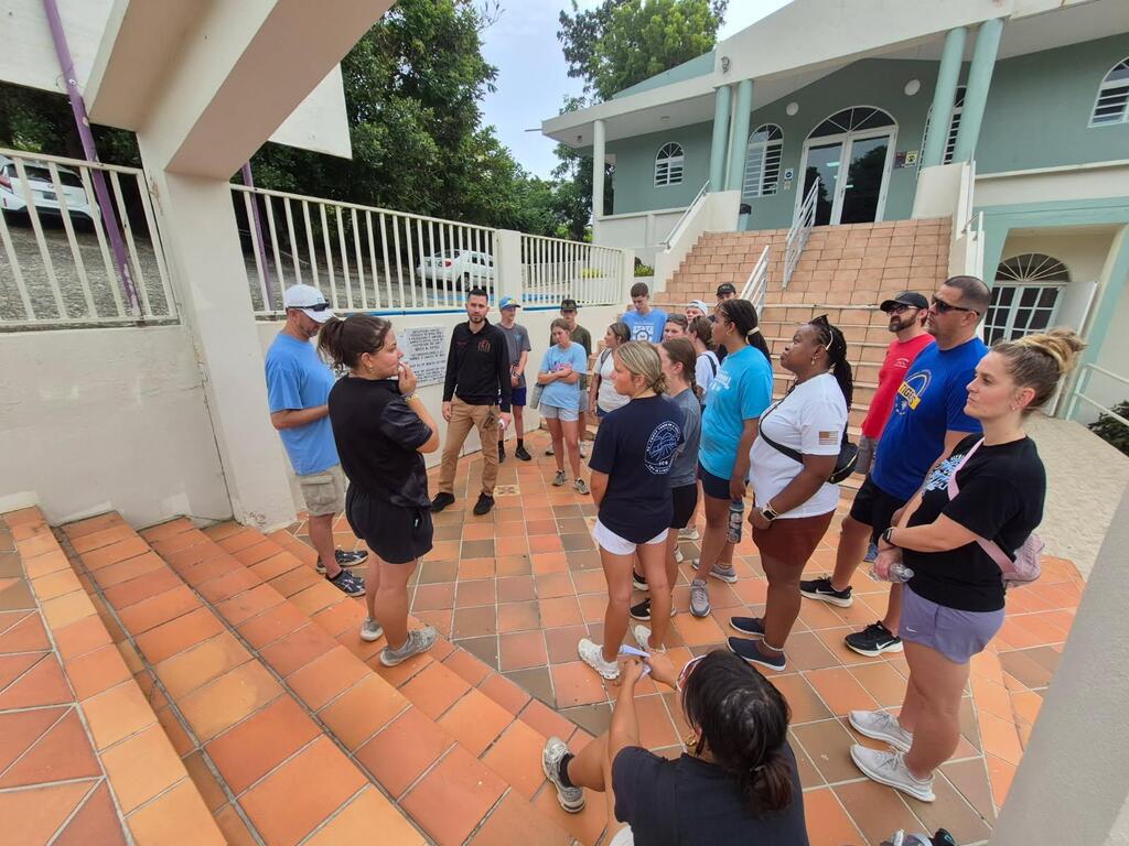 A group of people gather on a porch with tiled steps, some standing, others listening, near a house.
