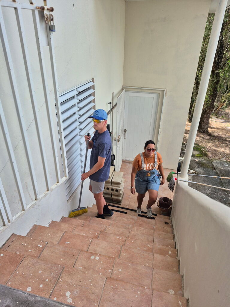 Two people are cleaning stairs with a broom. One person holds a broom, and the other holds a dustpan.