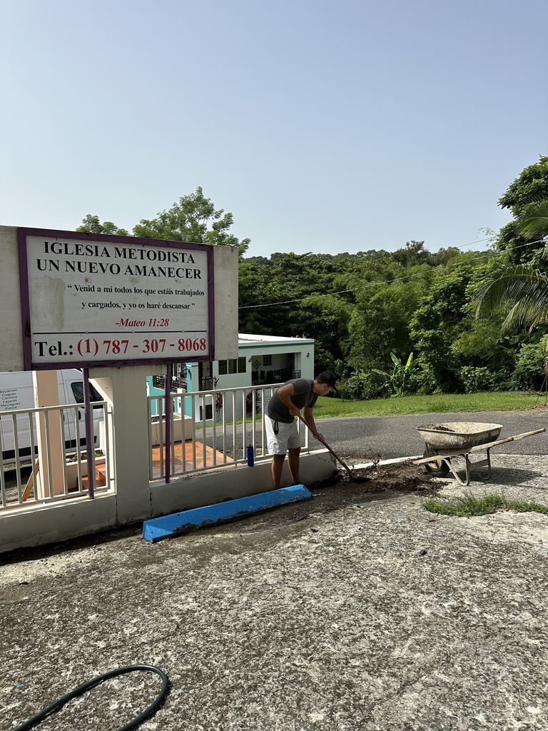 A person is working with a wheelbarrow on the road in front of a sign. Behind is a white van. Trees and greenery are visible in the background.