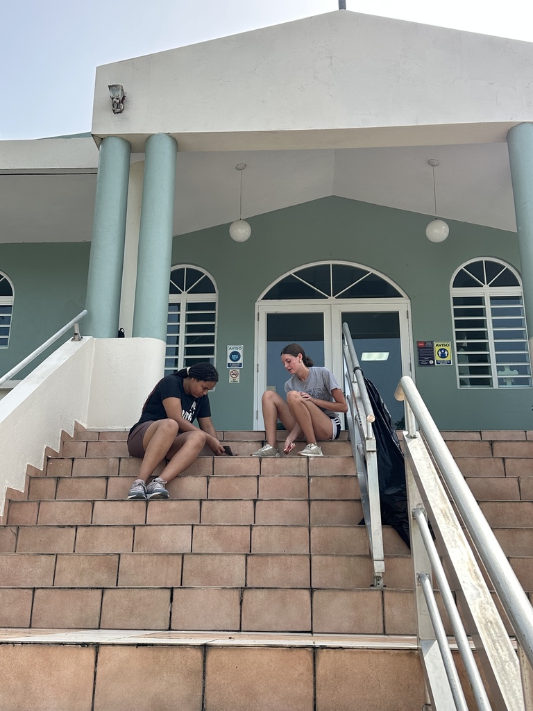 Two people sit on the steps outside of a building, one with a backpack, under clear skies.