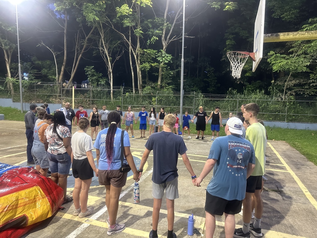 Several people are standing in a circle holding hands outside near a basketball court at night.