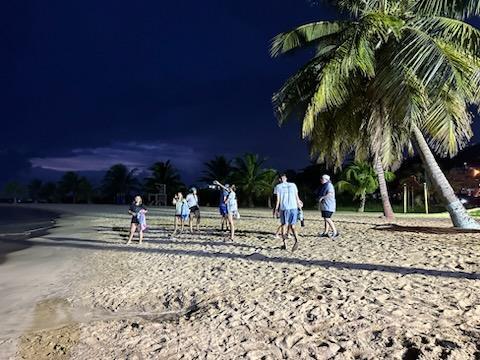A group of people in matching blue outfits walk on a sandy beach at night, with palm trees visible.