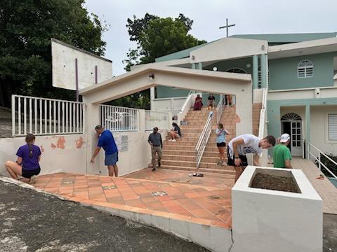 People in shorts and t-shirts working on a white and blue building with a basketball hoop, stairs, and trees.
