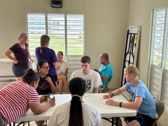 People seated around a table in a room with windows and blinds. Some individuals stand near the walls.