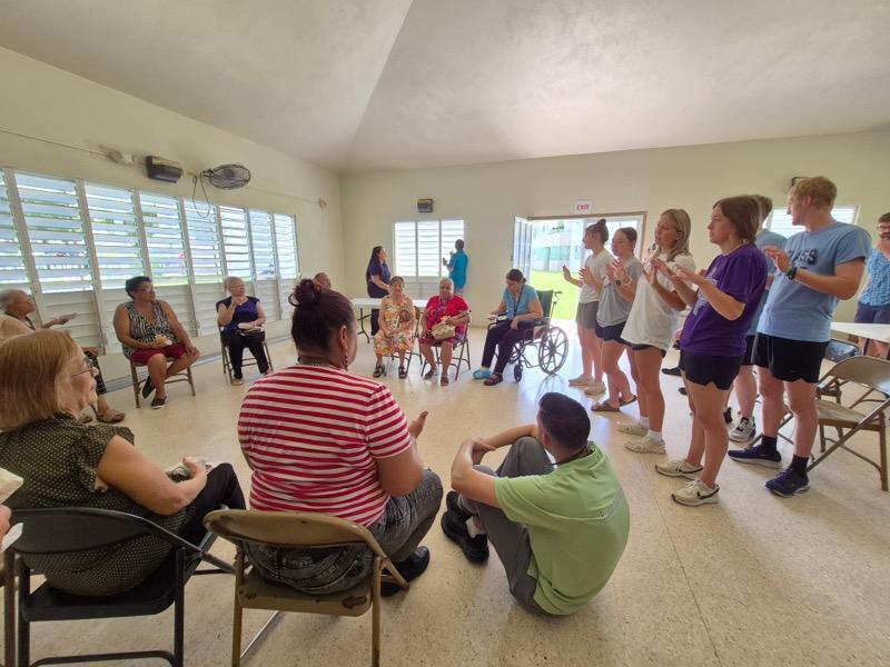 Group of people in a room, some seated, others standing. A few people in wheelchairs. One individual clapping hands.