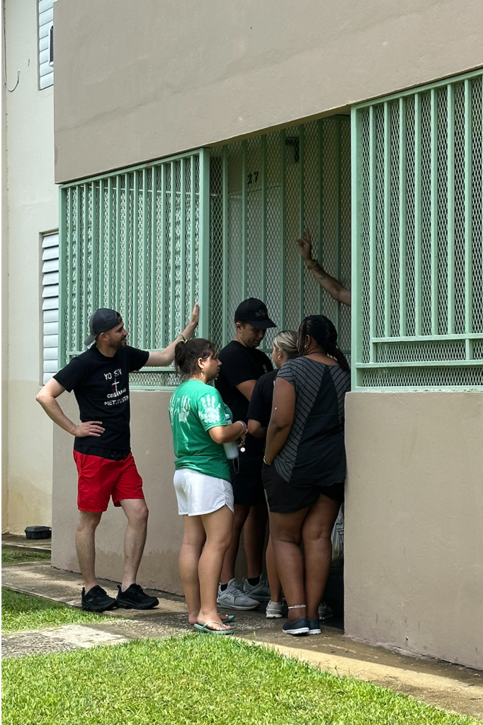 Five people gather outside a building, two standing and three inside a doorway, one holding the bars.
