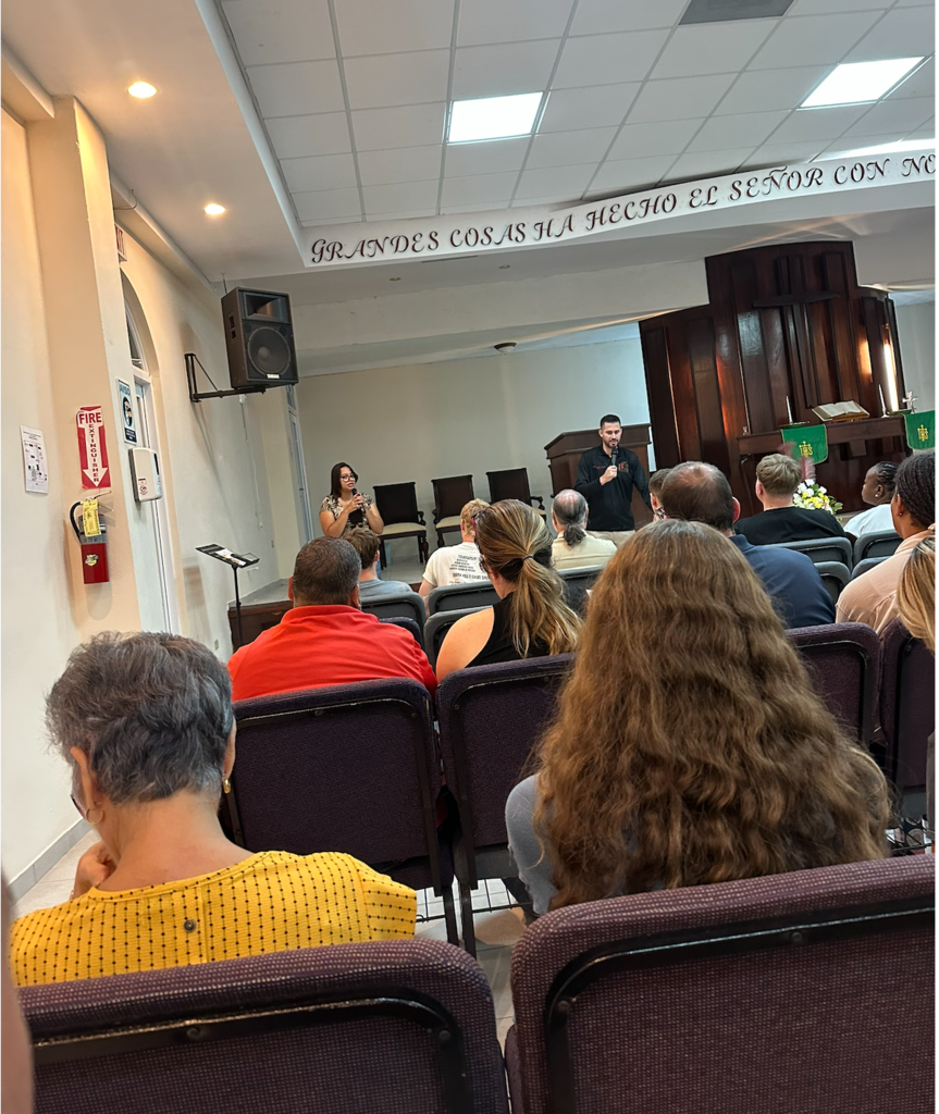 Group of people sit in chairs facing a stage with a speaker, a banner above, and a fire extinguisher on the wall.