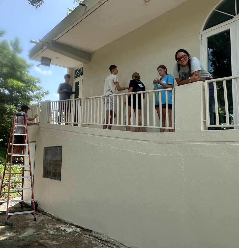People on the balcony and ground outside a house, with one person standing on a ladder.