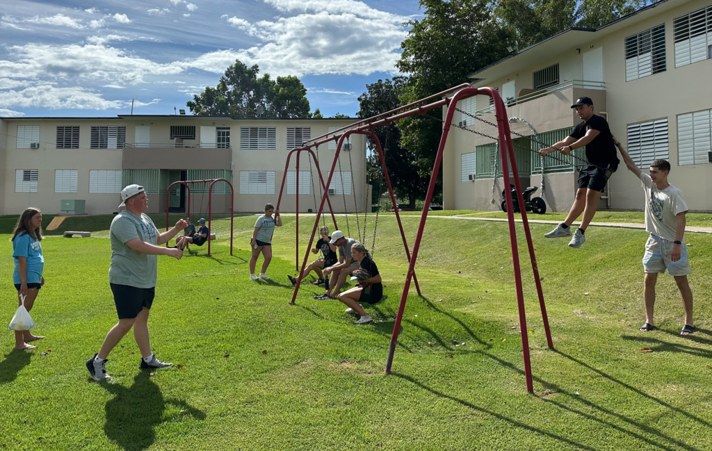 A group of people are on a grassy lawn. Some are using playground swings and others are walking. An apartment building is in the background.