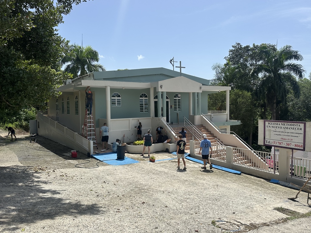 A building with stairs, a blue tarp, and people in a dirt area surrounded by trees.