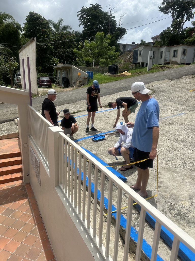 People are working on a project by a house on a street. They are standing and kneeling on the sand.