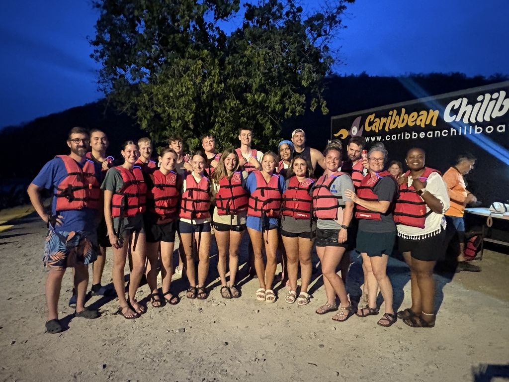Group of people wearing life jackets posing for a photo in front of a Caribbean Chillin banner.