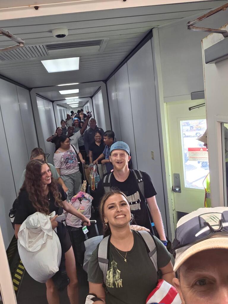 People in a hallway, some carrying bags. Ceiling lights and a door. A person takes a selfie.