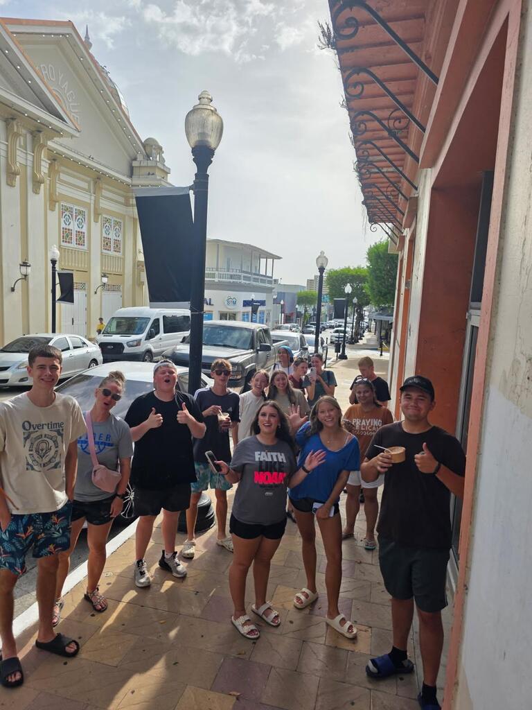 A group of people in summer clothing standing on a sidewalk with street lamps and parked cars.