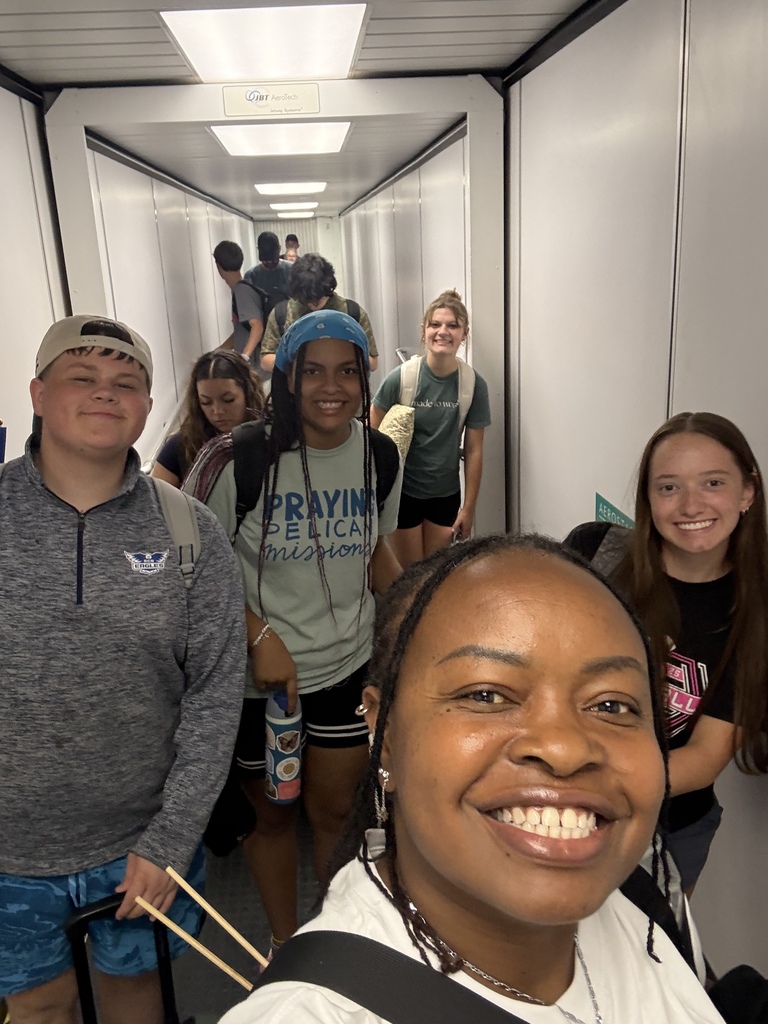A group of people walk through a hallway. They wear casual clothing and some have backpacks. One person holds chopsticks.