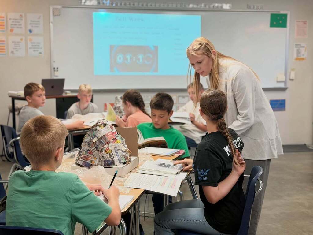 A teacher in white assists students seated at desks in a classroom with a projector screen displaying a red logo.