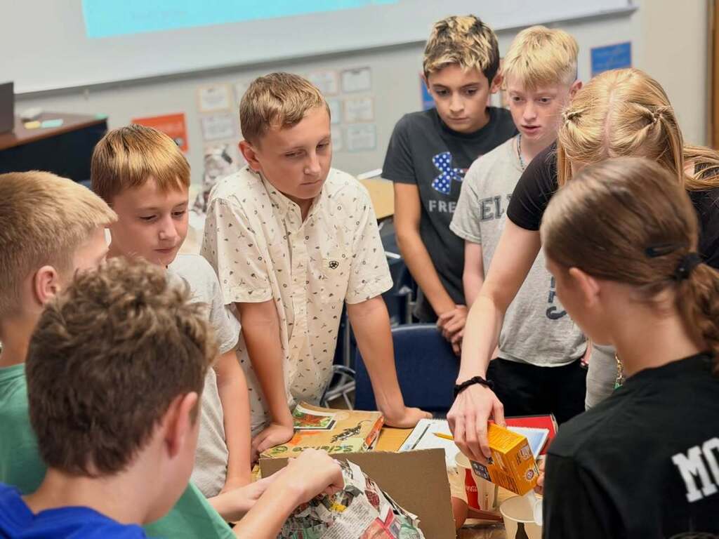 Classroom with children examining a box of food products while a teacher assists them.