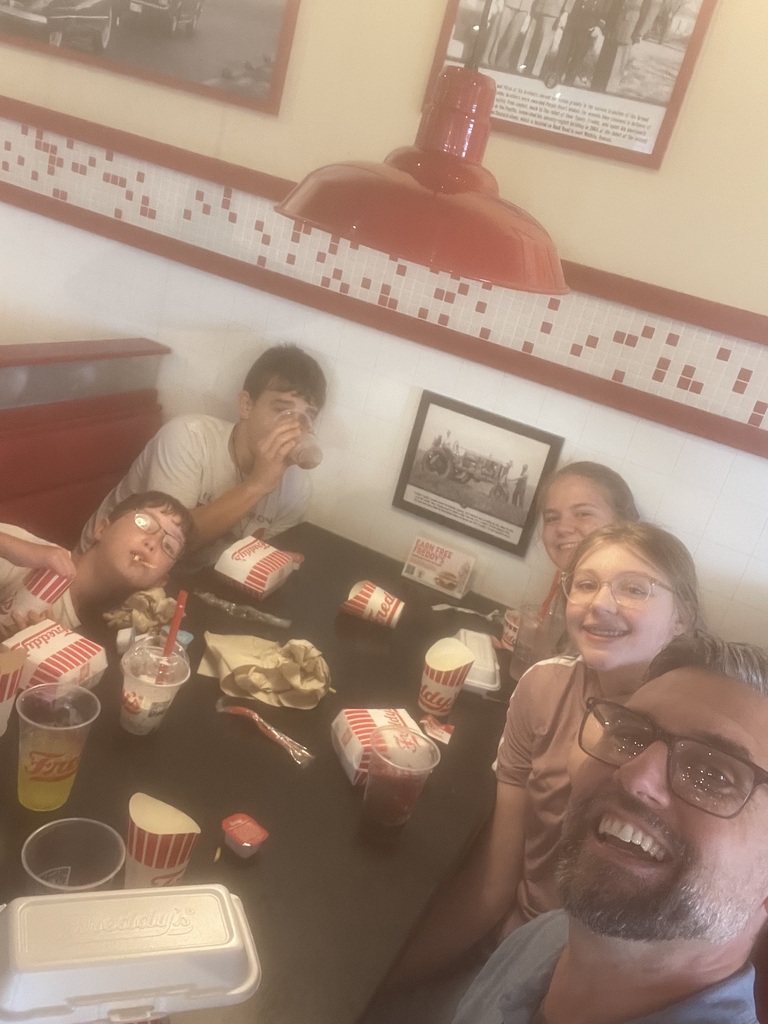 A group of people sits around a table at a restaurant, smiling. The table has food, cups, and napkins.