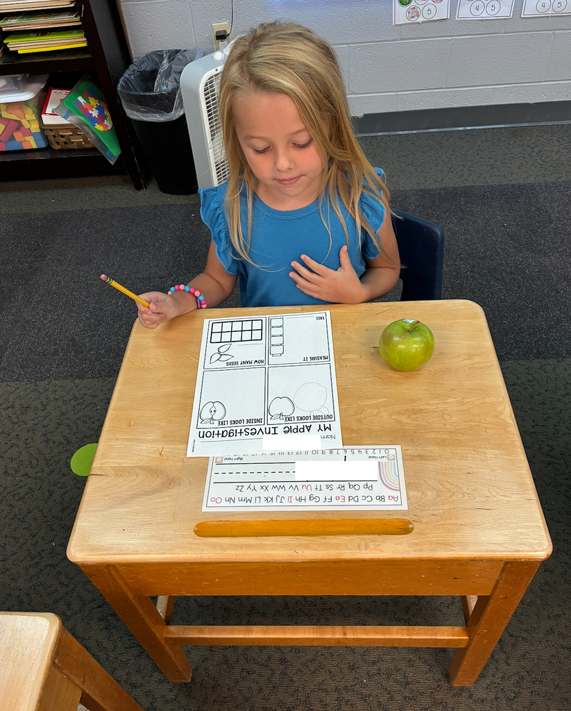 A young girl sits at a desk, holding a pencil. Papers are on the desk, and an apple is nearby.