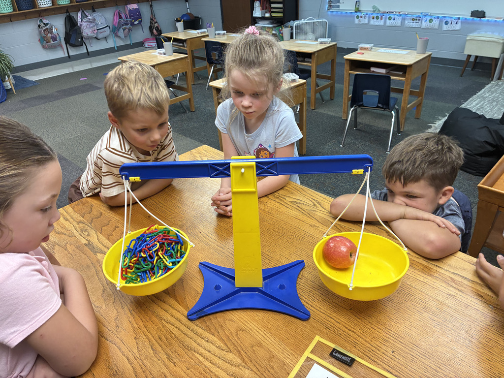 Four children sit around a table with a balance scale in a classroom. The scale has two bowls, one with colorful objects and one with an apple.