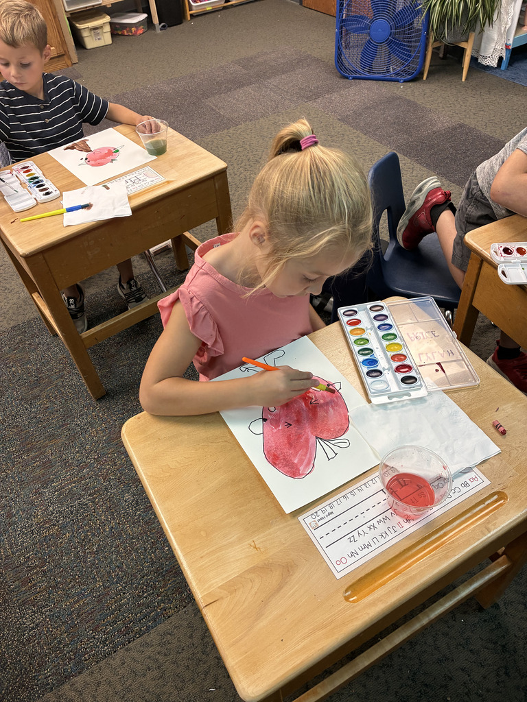 A girl painting a butterfly on paper at a desk with a palette of paints. Two other children are seated at nearby desks.