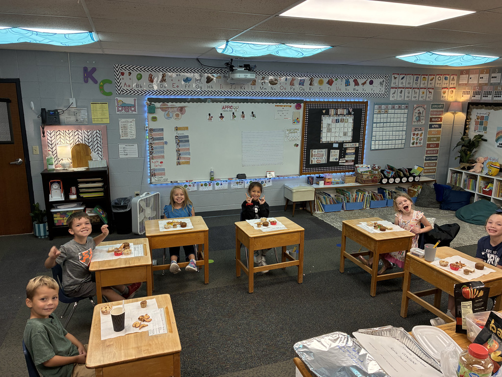 A classroom with children seated at desks. Desks have food on them. A whiteboard is mounted on the wall behind them.